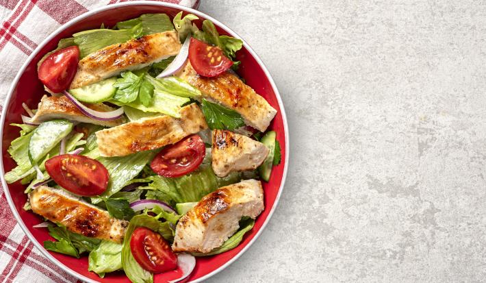 Fotografia vista de cima de um salada de folhas pepino, tomate, frango, cebola roxa e salsinha com um molho de maracujá e mel. A salada está dentro de um recipiente fundo vermelho, apoiado sobre e metade de um pano branco com linhas vermelhas.