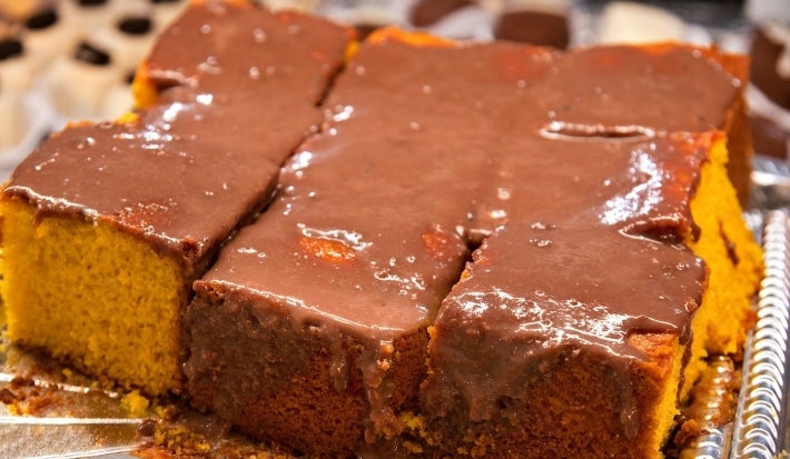 Bolo de cenoura cortado em pedaços quadrados, coberto com uma camada generosa de calda de chocolate brilhante. Os pedaços estão organizados sobre uma bandeja metálica.