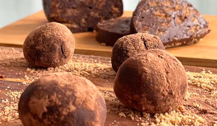 A imagem mostra quatro bolinhas de chocolate em formato de trufa sobre uma tábua de madeira. Ao fundo, uma massa de chocolate em formato cilindrico está cortada em fatias.