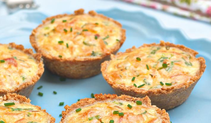 Fotografia em tons de azul em uma mesa de madeira azul com um prato redondo raso azul e alguns mini quiches de batata e salmão.