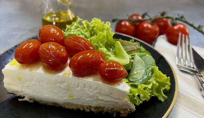 Foto da receita de Cheesecake Salgado. Observa-se uma fatia da torta com tomatinhos confit em cima em um prato preto servido com uma salada de alface.