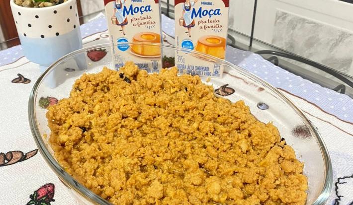 Foto da receita de ambrosia em tom amarelo, numa travessa de vidro, sobre uma mesa com toalha e nela há duas caixas do produto de Moça e uma vasinho com uma planta em tom verde