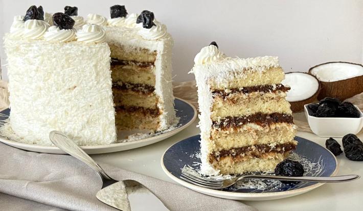 Fotografia em tons de branco com um bolo cortado ao centro. Ao lado existe uma fatia desse mesmo bolo, ele possui camadas de creme de coco e ameixa e está decorado com chantilly e coco ralado. Ao fundo existe algumas ameixas e um coco inteiro.