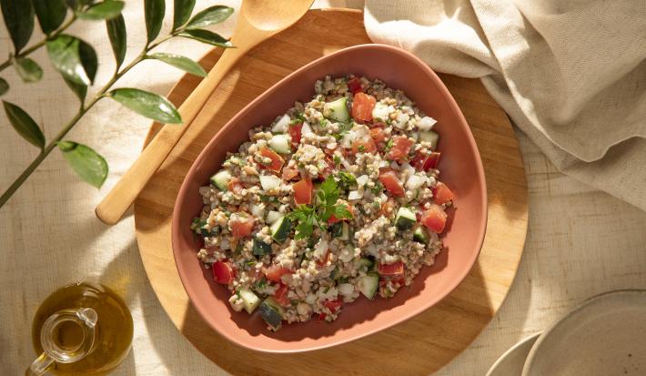Foto da receita de tabule de aveia corte rústico servida em um prato fundo rosa salmão sobre uma tábua de madeira circular com um garfo de madeira ao lado e um pano de linho