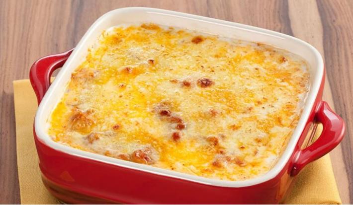 Fotografia em tons de vermelho em uma bancada de madeira com um pano amarelo e um recipiente quadrado com alças laterais vermelho e o frango gratinado dentro dele.