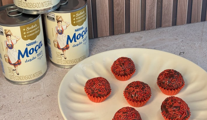 Docinhos de chocolate com granulado vermelho em forminhas vermelhas, servidos em prato claro com Leite Moça ao fundo.