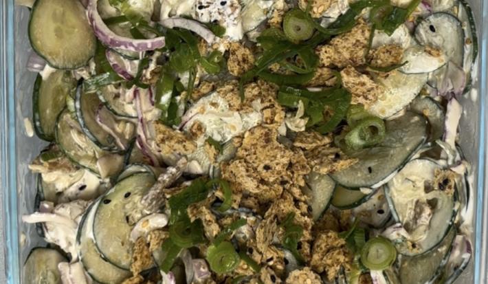 Fotografia em tons de verde com uma travessa de vidro ao centro. Dentro da travessa existe uma salada de pepino com cebola roxa e cream cheese