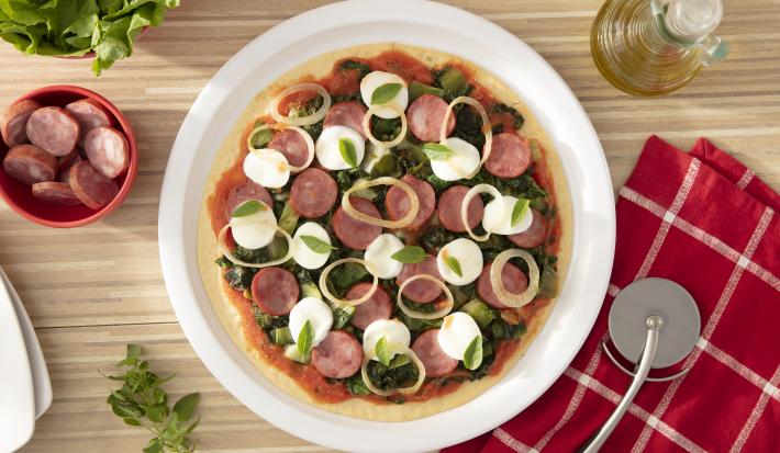 Imagem vista de cima de uma pizza redonda sobre um prato branco com calabresa, escarola, cebola, muçarela de búfala e manjericão.