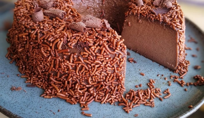 Fotografia de um prato raso, grande, de vidro azul suspenso com o brigadeirão de café sobre ele. O brigadeirão está com um pedaço grande cortado, mostrando a textura da sobremesa, que está finalizada com granulado e raspas de chocolate.