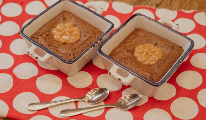 Fotografia em tons de marrom e vermelho de uma bancada de madeira sobre ela um paninho vermelho com bolinhas brancas com duas colheres se sobremesa. Sobre ele dois recipientes quadrados e brancos, dentro o brownie e o MOÇA doce de leite.