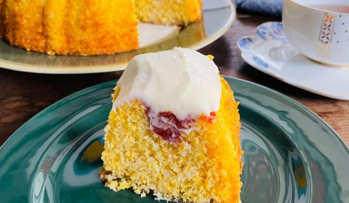 Fotografia em tons de verde e amarelo com um prato verde ao centro. Em cima do prato existe uma fatia de bolo de fubá com cobertura de goiabada e cream cheese. Ao fundo existe o bolo inteiro com uma fatia cortada