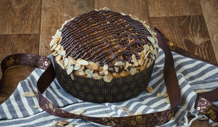 Imagem da receita de Colomba Pascal, decorada com tiras de chocolate e amêndoas em lascas, sobre uma bancada decorada com um tecido azul e branco, com uma fita de páscoa decorativa