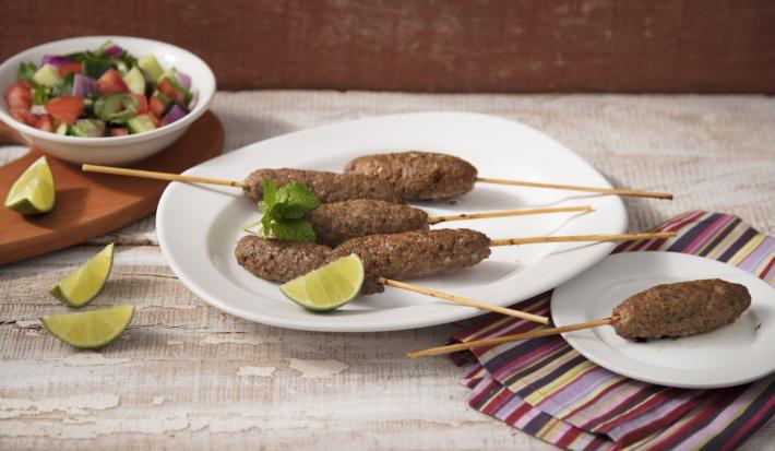 fotografia em tons de bege, marrom e branco de uma bancada bege vista de frente, contém um prato branco redondo com palitos de bolinhos de carne moída e fatias de limão, ao lado um pano com um prato branco e redondo com um espetinho com a kafta