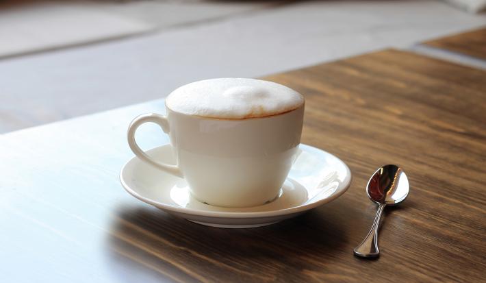 fotografia em tons de marrom e branco tirada de uma bancada marrom. Contém um pratinho branco com uma xicara de café branca, e dentro contém a bebida Cappuccino.