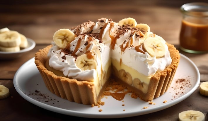 Em cima de uma mesa, prato com fatia de banoffee com massa de biscoitos crocante, bananas em rodelas, doce de leite, decorado com chantilly cremoso, banana em rodelas e cacau em pó.