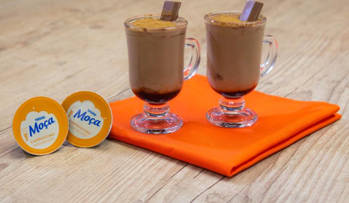 Fotografia em tons de laranja em uma bancada de madeira de cor marrom. Ao centro, um pano laranja contendo duas taças com as bebidas. Ao lado das taças há duas cápsulas de Nescafé.