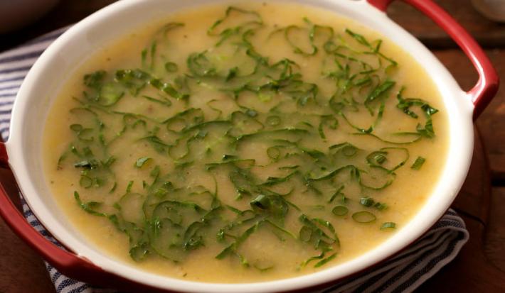 Fotografia em tons de marrom e branco de uma bancada de madeira com paninho listrado azul e branco sobre ele uma panela com sopa de mandioca com couve.
