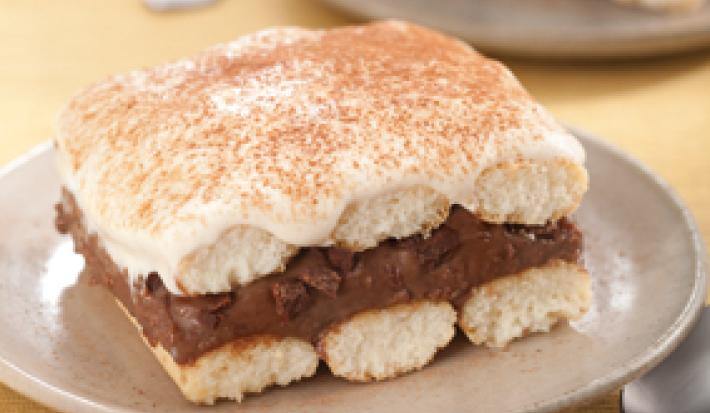Fotografia em tons de bege em uma mesa de madeira com uma toalha amarela e um prato bege redondo raso ao centro com um pedaço de pavê de chocolate.