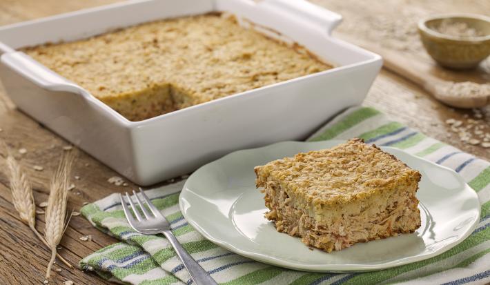 Fotografia em tons de branco e dourado, refratário branco com torta de frango, prato branco com uma porção de torta de frango com um garfo ao lado e sobre guardanapo com listras verdes, azuis e brancas, colher e potinho com aveia ao fundo.
