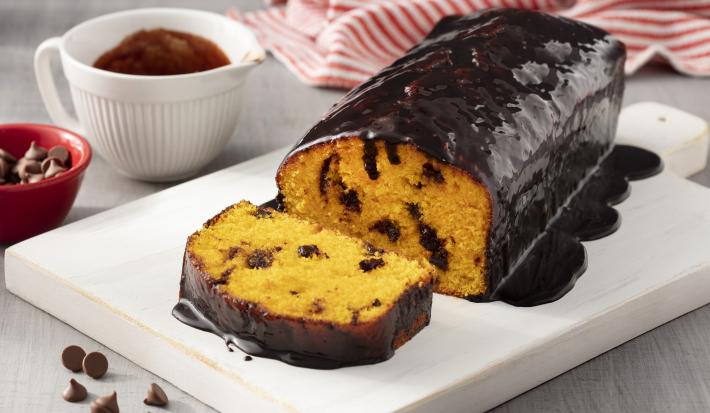 Fotografia em tons de branco e vermelho de uma bancada branca branca com um prato retangular branco, sobre ele um bolo de cenoura. Ao fundo um paninho listrado vermelho e branco e um recipiente redondo branco com calda de chocolate.
