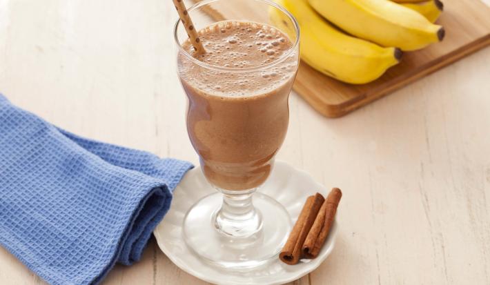 Fotografia em tons de marrom em uma bancada de madeira clara com um pano azul ao lado e ao centro um pratinho branco com uma taça de vidro e o smoothie de cacau e banana dentro. Ao fundo, um cacho de banana apoiado em uma tábua de madeira.