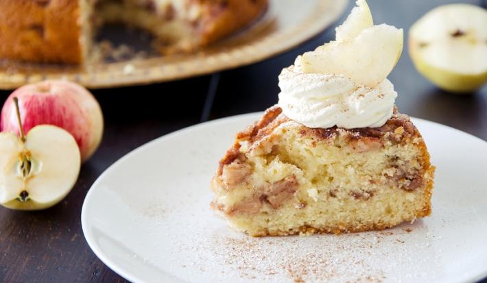Fotografia em tons de branco em uma bancada de madeira de cor preta. Ao centro, um prato branco contendo a torta e ao fundo, uma maçã cortada ao meio