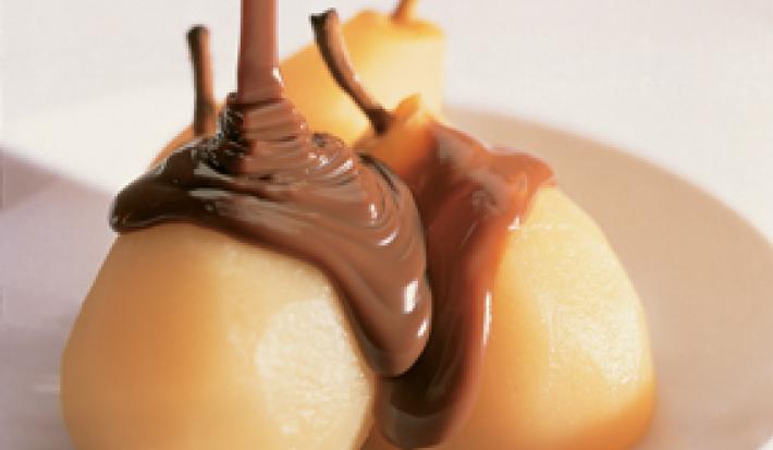 Fotografia em tons de amarelo em uma bancada de madeira branca, um prato redondo raso branco com as peras e calda de chocolate em cima dele.