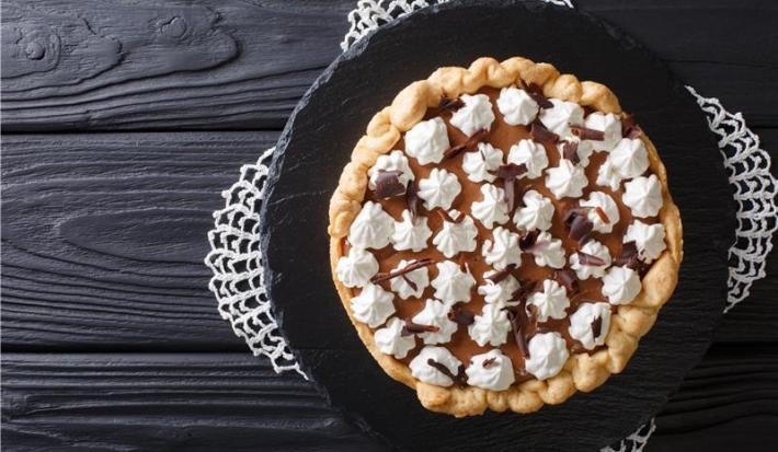 Fotografia vista de cima de uma torta redonda com chantilly em bicos com raspas de chocolate. A torta está sobre um apoio preto, o qual está apoiado em uma mesa de madeira preta.