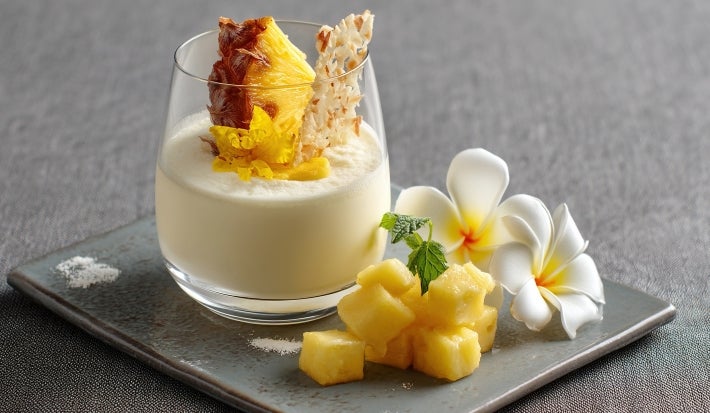Imagem de um copo servido com um creme cremoso de abacaxi, decorado com uma fatia de abacaxi em meia-lua e lascas de coco.