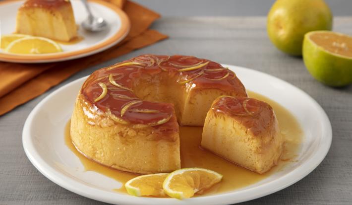 Receita de Pudim de Laranja. Observa-se um prato grande branco contendo o pudim com calda e raspas. Atrás, um prato de sobremesa com uma fatia do pudim e uma colherinha. Ao lado, laranjas decoram a foto.