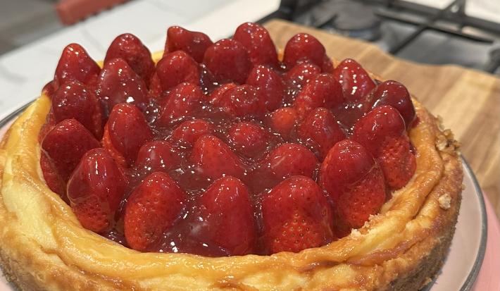 Fotografia em tons claros de uma receita de cheesecake com morango em um prato raso de tom pastel sobre uma tábua de madeira.