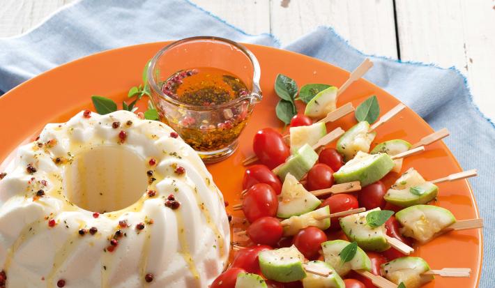 Fotografia em tons de branco, laranja e azul com prato laranja contendo mini espetinhos de vegetais, potinho com molho e uma terrine branca, sobre guardanapo azul e bancada na cor branca.