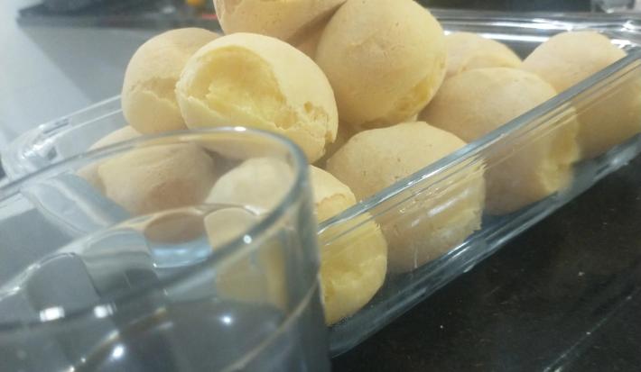 Fotografia em tons de branco em uma bancada de madeira de cor preta. Ao centro, uma travessa de vidro contendo os pães de queijo.