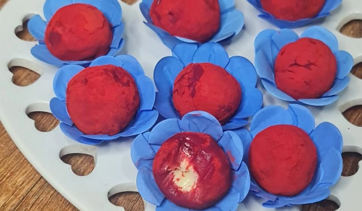 Foto em tons de vermelho da receita de brigadeiro red velvet servida em diversas porções sobre embalagens azuis, que se dispõem em cima de uma base em formato de coração branca sobre uma mesa de madeira