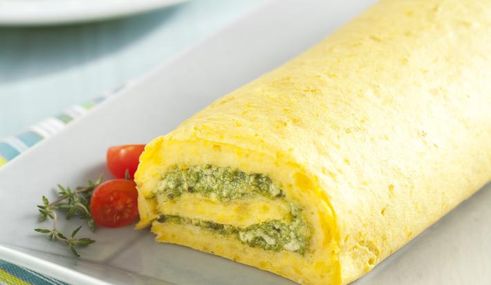 Fotografia em tons amarelados com um paninho listrado em azul, cinza e amarelo e um prato retangular com o rocambole de espinafre, mandioquinha e tofu em cima. Ao fundo, um prato com um pedaço do rocambole e um garfo azul ao lado.