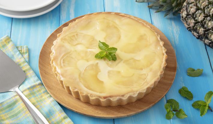Receita de Torta de Abacaxi ao Creme de Ricota. Observa-se uma torta inteira redonda decorada com hortelã e abacaxi sobre uma tábua redonda de madeira em uma superfície de madeira azul clara. Ao lado esquerdo, uma espátula prateada.