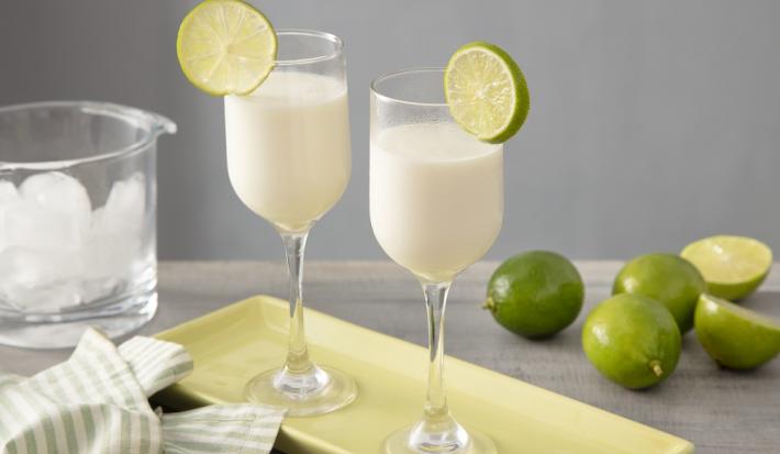 Foto em tons claros de verde da receita de nevada com leite MOÇA servida em duas taças largas e altas sobre uma base de porcelana verde, ao lado há um pano listrado nos tons da foto e ao fundo há limões e um ricipiente de vidro com gelo.