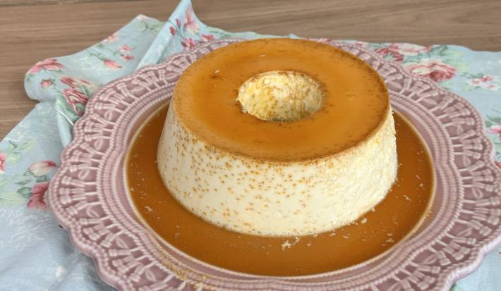 Fotografia em tons de rosa com um pudim de Leite MOÇA ao centro. Por cima do pudim existe uma calda de caramelo