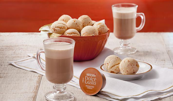 fotografia em tons de laranja e cinza de uma bancada cinza vista de cima, contém um pano branco, um recipiente redondo laranja com vários pães de queijo, duas xicaras transparentes com Mochaccino Canela, um prato redondo com dois pães de queijo.