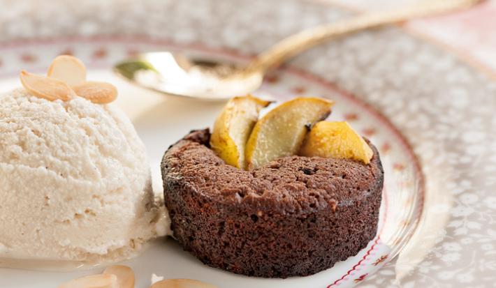 Fotografia em tons de salmão em uma bancada de madeira com uma toalha branca com detalhes salmão, um prato redondo de porcelana desenhado em cinza e rosa com uma bola de sorvete de creme e um mini bolinho de chocolate em cima dele.