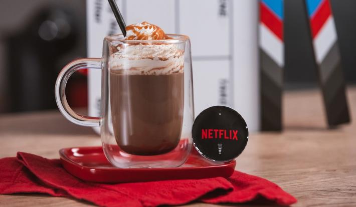 Foto em tons de vermelho e preto da receita de cappuccino netflix servida em um copo largo sobre uma base de porcelana vermelha e um pano vermelho em cima de uma mesa de madeira com uma cápsula de cappuccino netflix ao lado