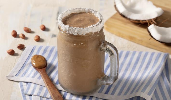 Pano listrado de azul claro e branco, com uma colher de madeira marrom com pasta de amendoim. Uma caneca transparente com o smoothie e borda de coco ralado.