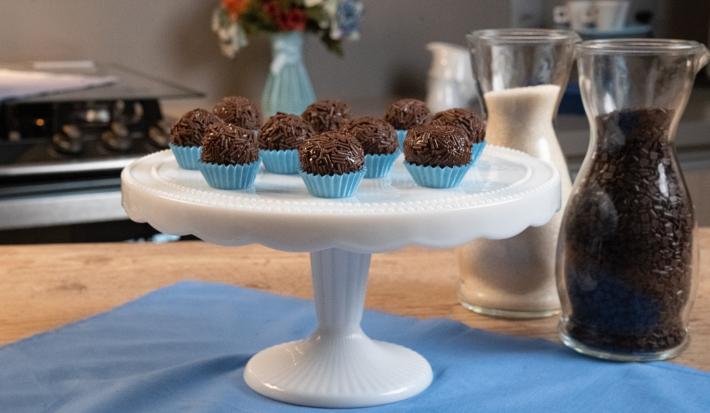 Fotografia de dez forminhas azuis com brigadeiros passados no granulado e apoiados em um enfeite para bolo branco e alto. Ao fundo tem um fogão, um vaso azul com flores, e ao lado do brigadeiro tem duas jarras decorativas de vidro com açúcar e cacau.