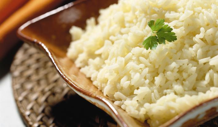 Fotografia em tons de marrom em uma mesa de madeira cinza com um descansa prato de palha marrom, um pano laranja e um recipiente retangular marrom com o arroz dentro.
