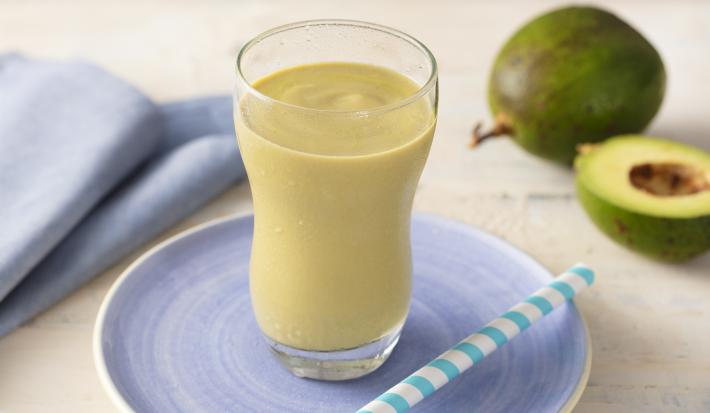 Fotografia em tons de azul e verde de uma bancada com um pratinho azul, um copo transparente com uma vitamina de abacate e um canudo. Ao fundo um paninho azul e um abacate.