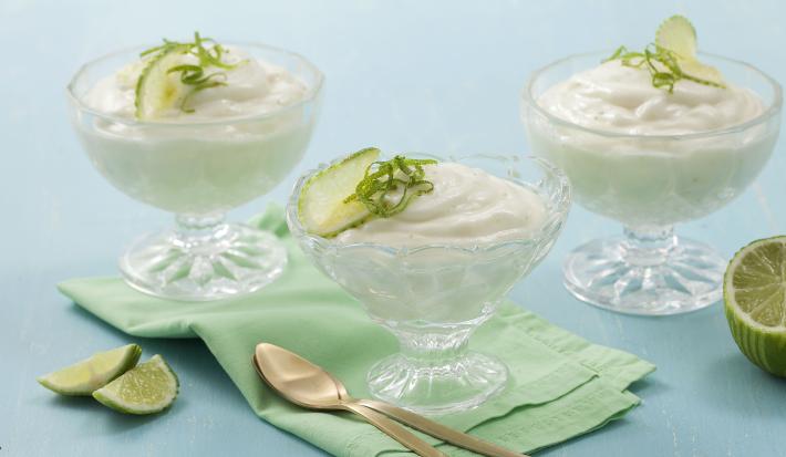 Fotografia em tons de azul e verde de uma bancada azul claro, com duas fatias de limão, um pano verde claro dobrado, duas colheres douradas, um limão pela metade e três taças transparentes com a mousse e raspas e rodelas de limão para decorar.