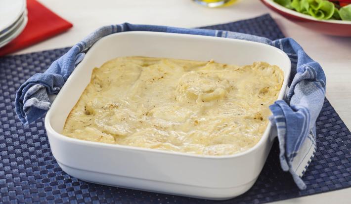 Fotografia em tons de azul e branco de uma bancada vista de cima. Contém um pano azul com um recipiente quadrado branco com alças e dentro as batatas gratinadas. Ao lado um pano azul.