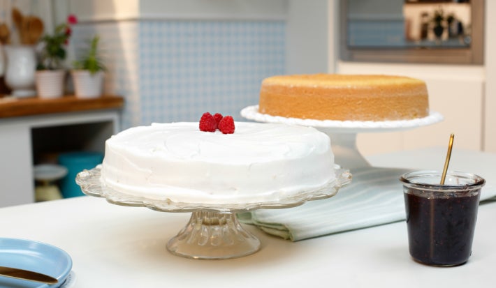 Imagem de dois bolos sobre boleiras: um coberto com chantilly e decorado com framboesas e um pão de ló simples.