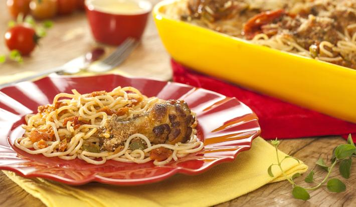 Fotografia em tons de amarelo e vermelho em uma bancada de madeira clara, panos amarelo e vermelho, um recipiente amarelo com o macarrão espaguete com asa de frango dentro dele, um prato vermelho com um pouco do macarrão.