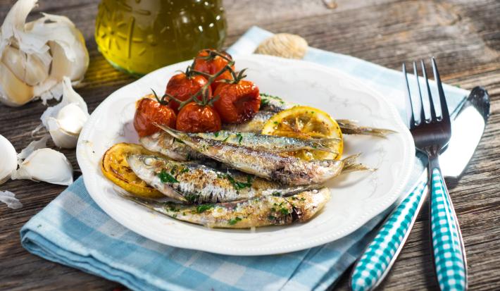 Fotografia em tons de branco com um prato branco e redondo ao centro. Em cima do prato existe sardinhas assadas com tomates e limão siciliano. Ao lado existe um garfo e uma faca de cor prateada.